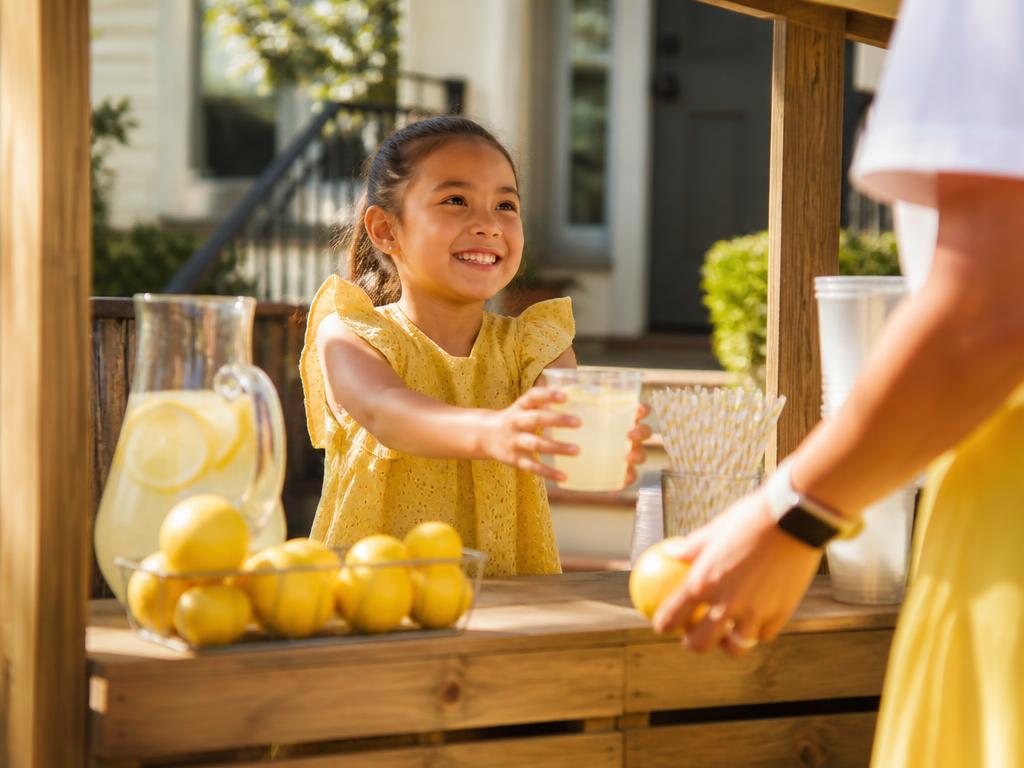 A young entrepreneur serving lemonade at her stand with a guardian helping
